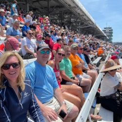 Crowd enjoying a sunny day at a motorsport race event, sitting in bleachers at a packed stadium.
