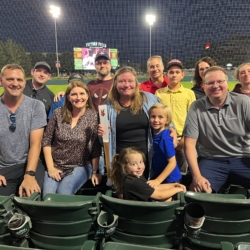 Group of people smiling at a night baseball game at Victory Field, with scoreboard in the background.