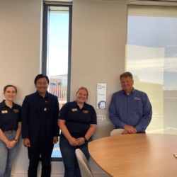 Four people smiling in a modern office setting, standing near a large round table by a window.