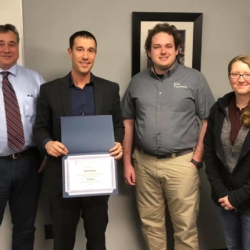Four people standing together, one holding a certificate, smiling in an office setting.