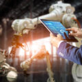 Factory worker using a tablet to control automated robotic arms in an industrial setting.